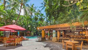 An outdoor seating area with wooden tables and chairs, a red umbrella, and a pool in the background. - 1 BR Suite City View, w/ AC, WiFi and Kitchen - Bamboo Waikīkī Hotel #504 (Honolulu)