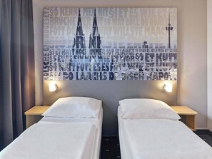 Une chambre d’hôtel avec deux lits simples, une grande œuvre d’art représentant un paysage urbain accrochée au mur, et deux tables de chevet avec des lampes.