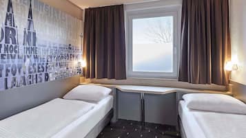 Une chambre d’hôtel avec deux lits simples, une fenêtre avec des rideaux, un bureau fixé au mur et une affiche représentant un paysage urbain.