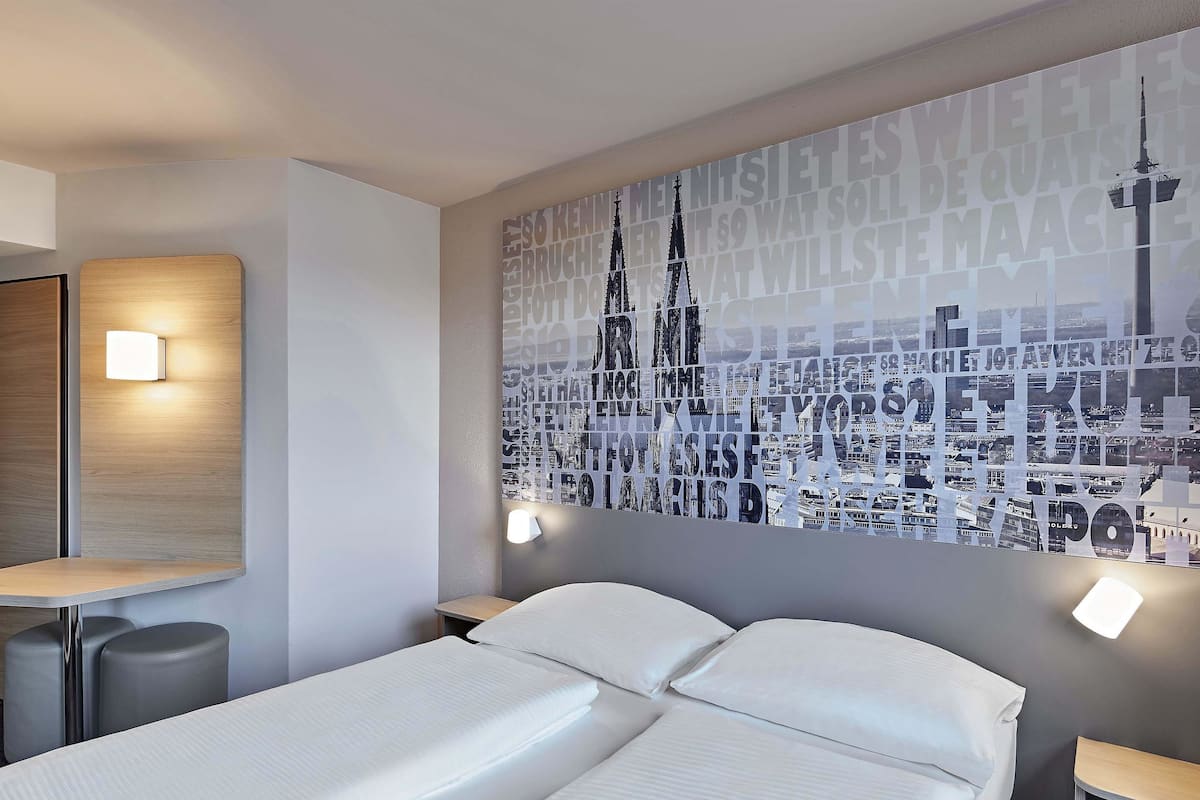 A hotel room with two beds, a large cityscape wall art, and a small table with a lamp.