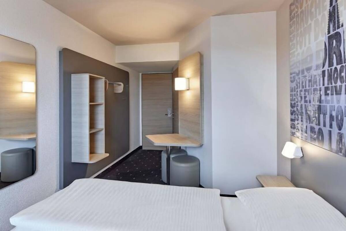 A hotel room with a bed, a small table, a wall-mounted shelf, and a door leading to a bathroom.