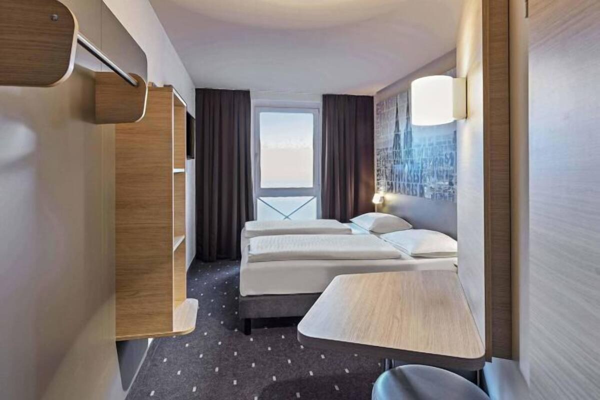 A hotel room with two beds, a desk, a wardrobe, and a cityscape view.