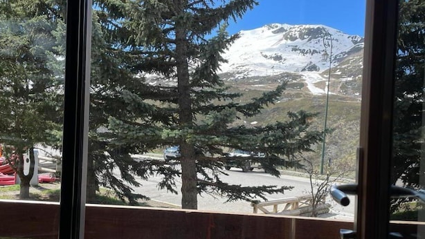 Une vue sur la montagne, visible depuis une fenêtre ornée d’une balustrade en bois, avec une table de pique-nique et un sommet enneigé en arrière-plan.