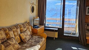 Une pièce chaleureuse avec un canapé à motifs, une petite table avec une lampe, un téléviseur et une grande fenêtre donnant sur un paysage enneigé.
