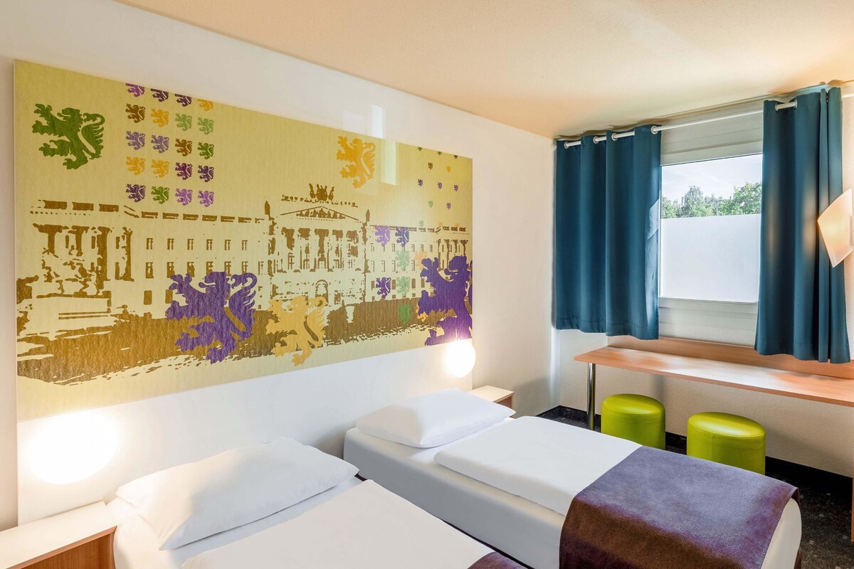 Habitación de hotel con dos camas, un mural grande en la pared, un escritorio con dos taburetes verdes y una ventana con cortinas azules.