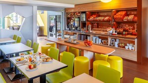 A modern breakfast area with yellow chairs, a counter with breakfast items, and a view of the street. - B&B Hotel Braunschweig-Nord (Braunschweig)