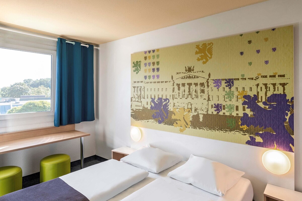 Habitación de hotel con cama, dos taburetes verdes, un escritorio y un mural grande en la pared.