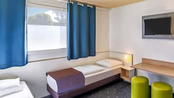 A hotel room with a bed, a bedside table with a lamp, a TV mounted on the wall, and two green ottomans.