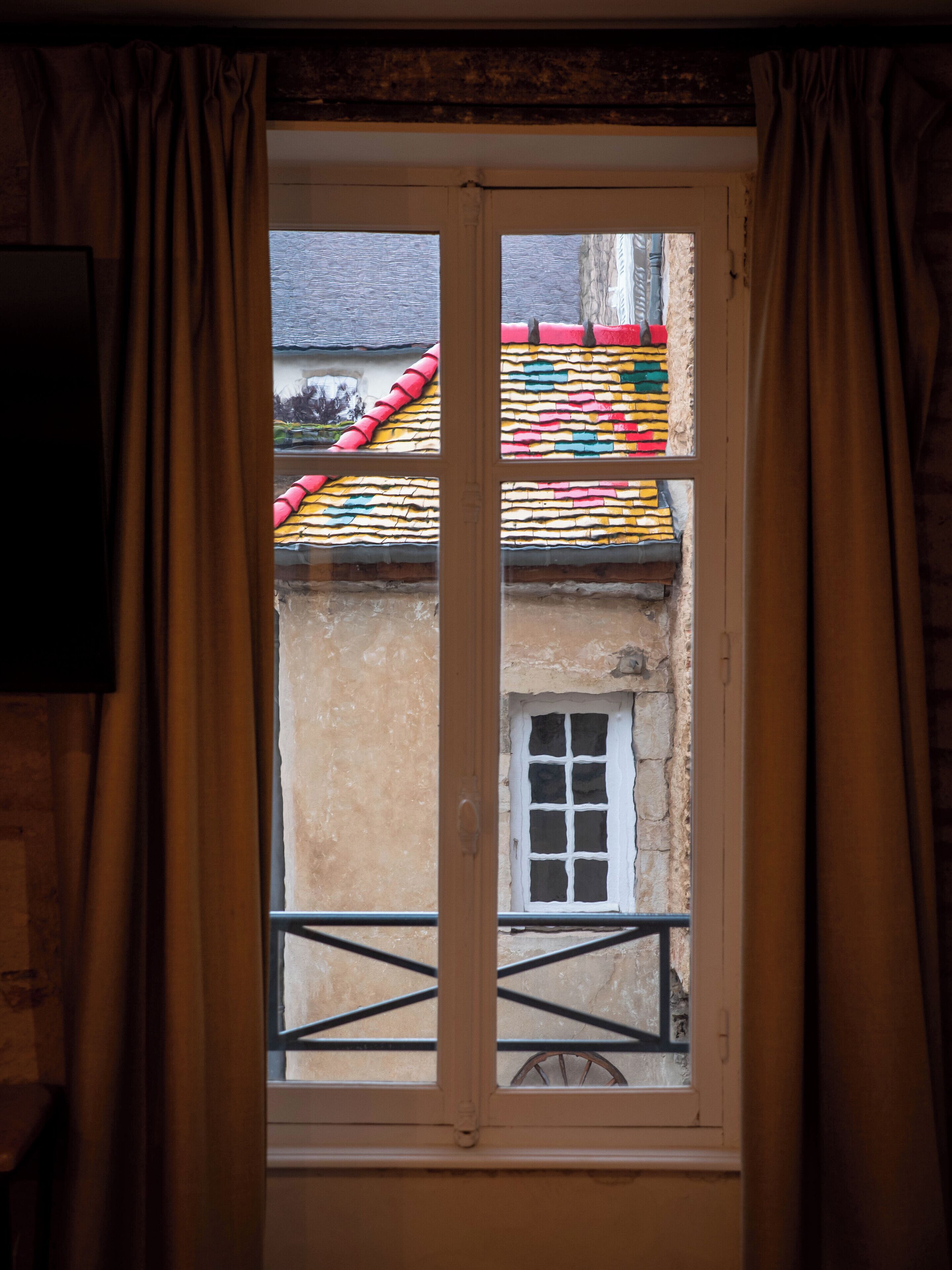 A window with a view of a building's colorful roof and a small window with white shutters.