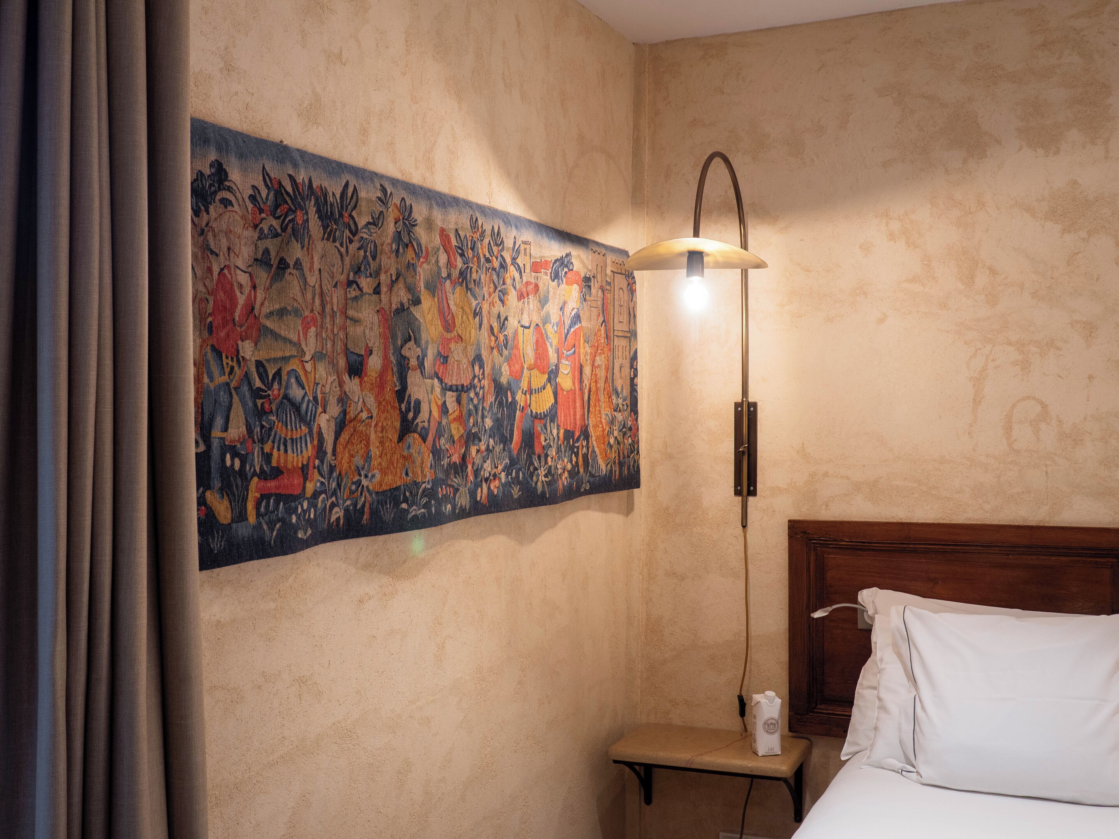 Une chambre d’hôtel avec une grande tapisserie au mur, une table de chevet avec une lampe et un lit recouvert de draps blancs.