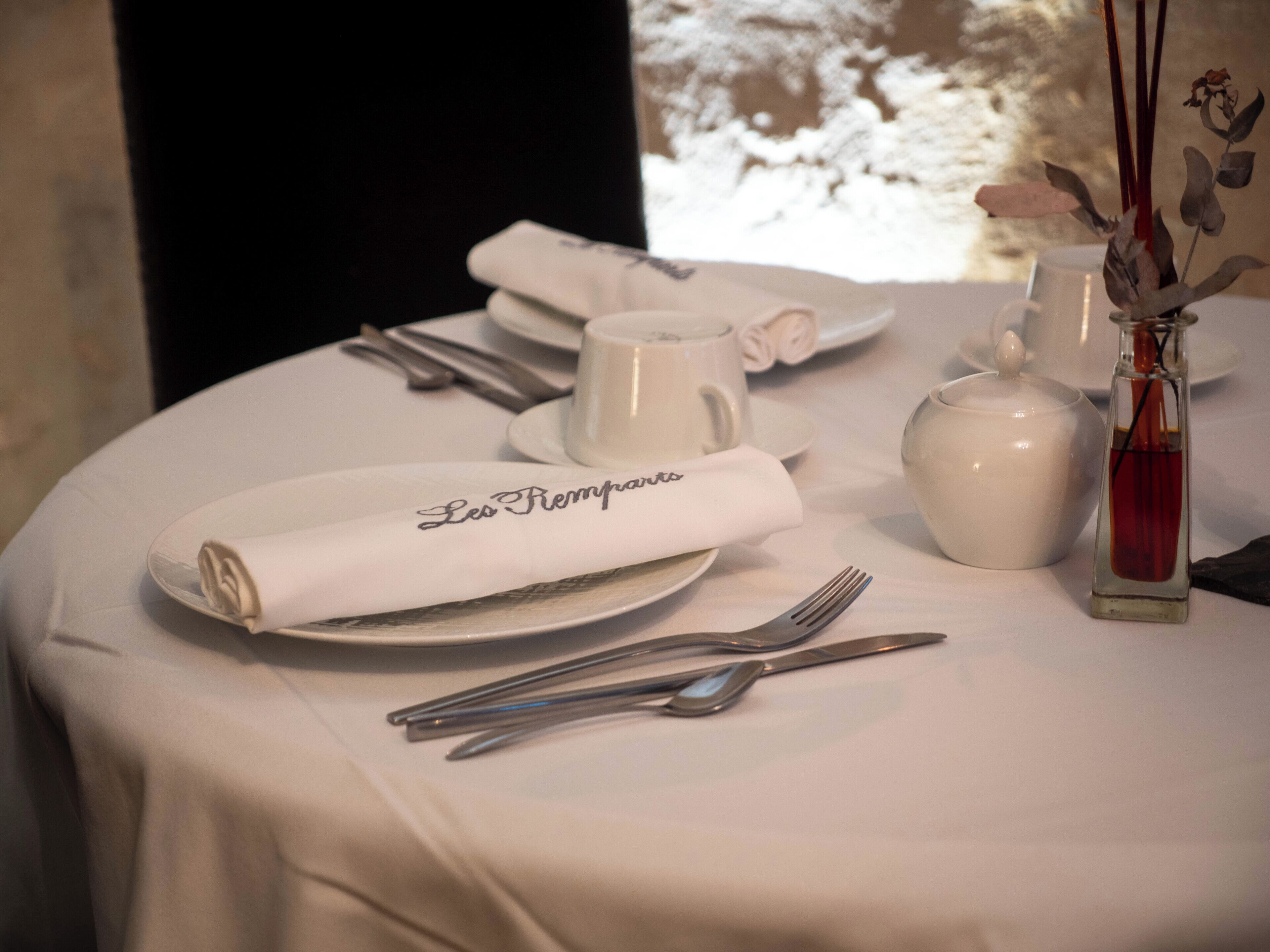 A table set with white tablecloth, plates, cutlery, a folded napkin, a teapot, and a vase with dried flowers.
