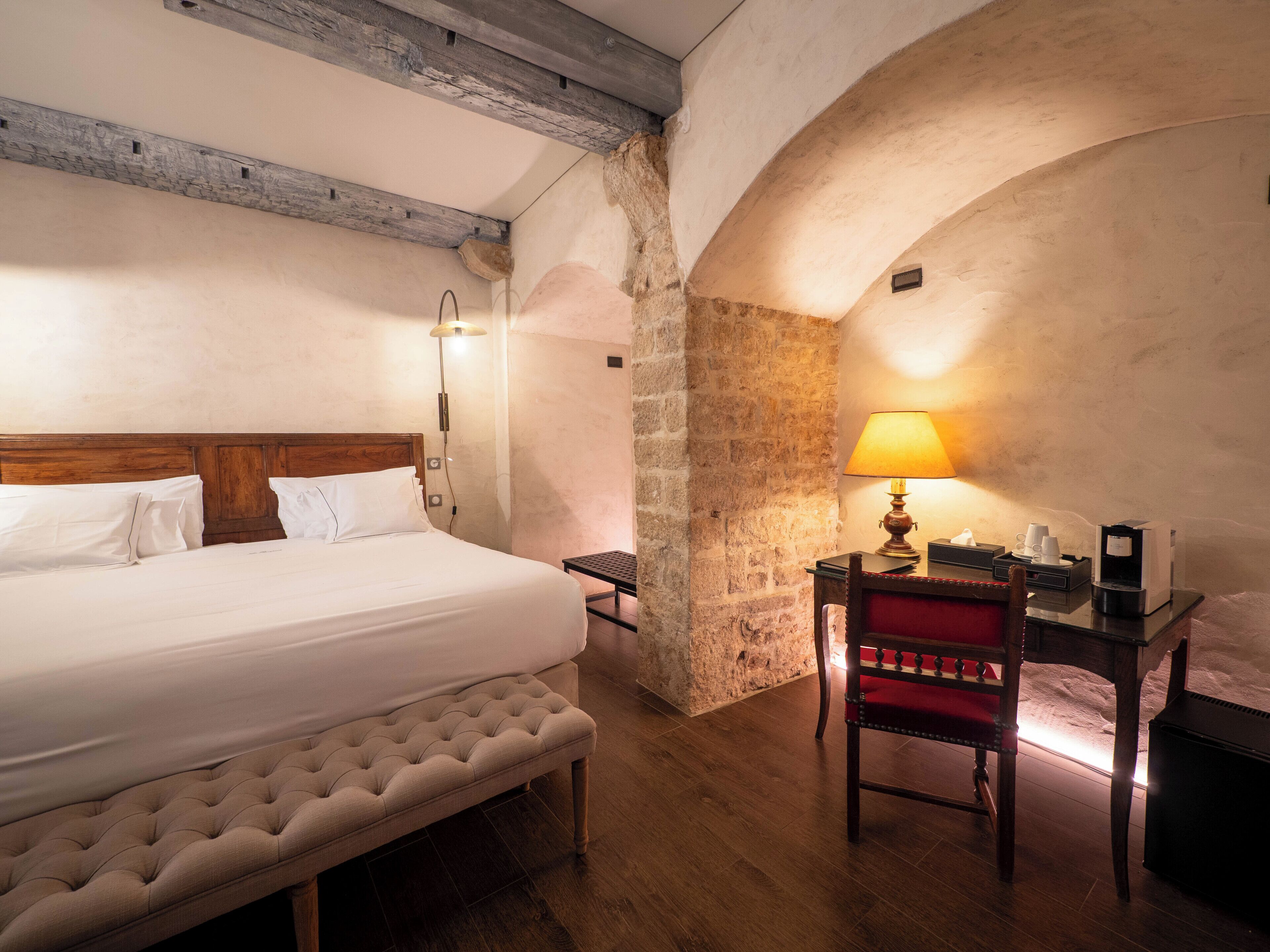 A hotel room with a large bed, a wooden bench, a desk with a lamp, and a coffee machine.