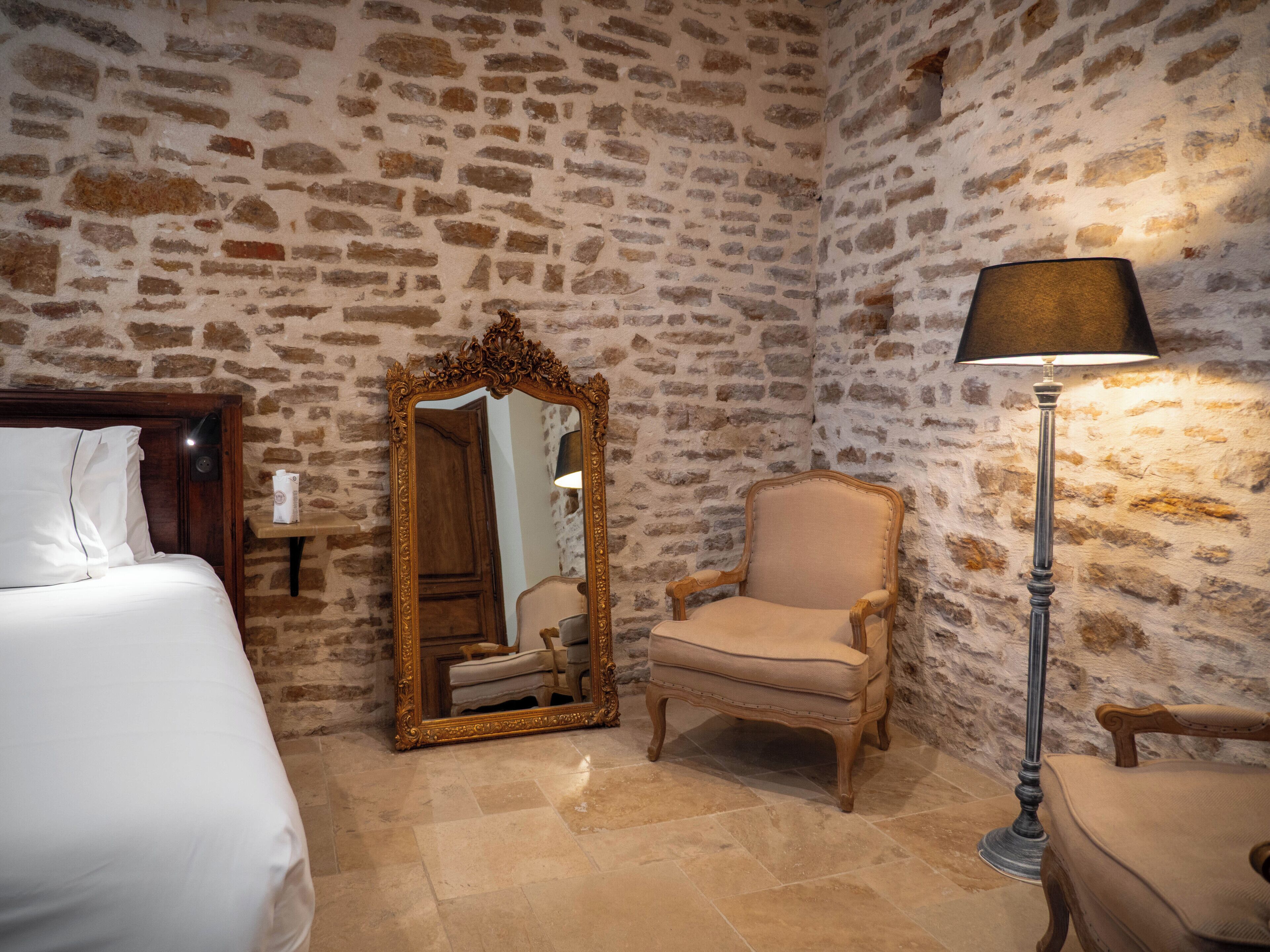 A room with a stone wall, a wooden bed, a chair, a mirror, and a floor lamp.