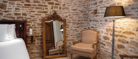 A room with a stone wall, a wooden bed, a chair, a mirror, and a floor lamp.