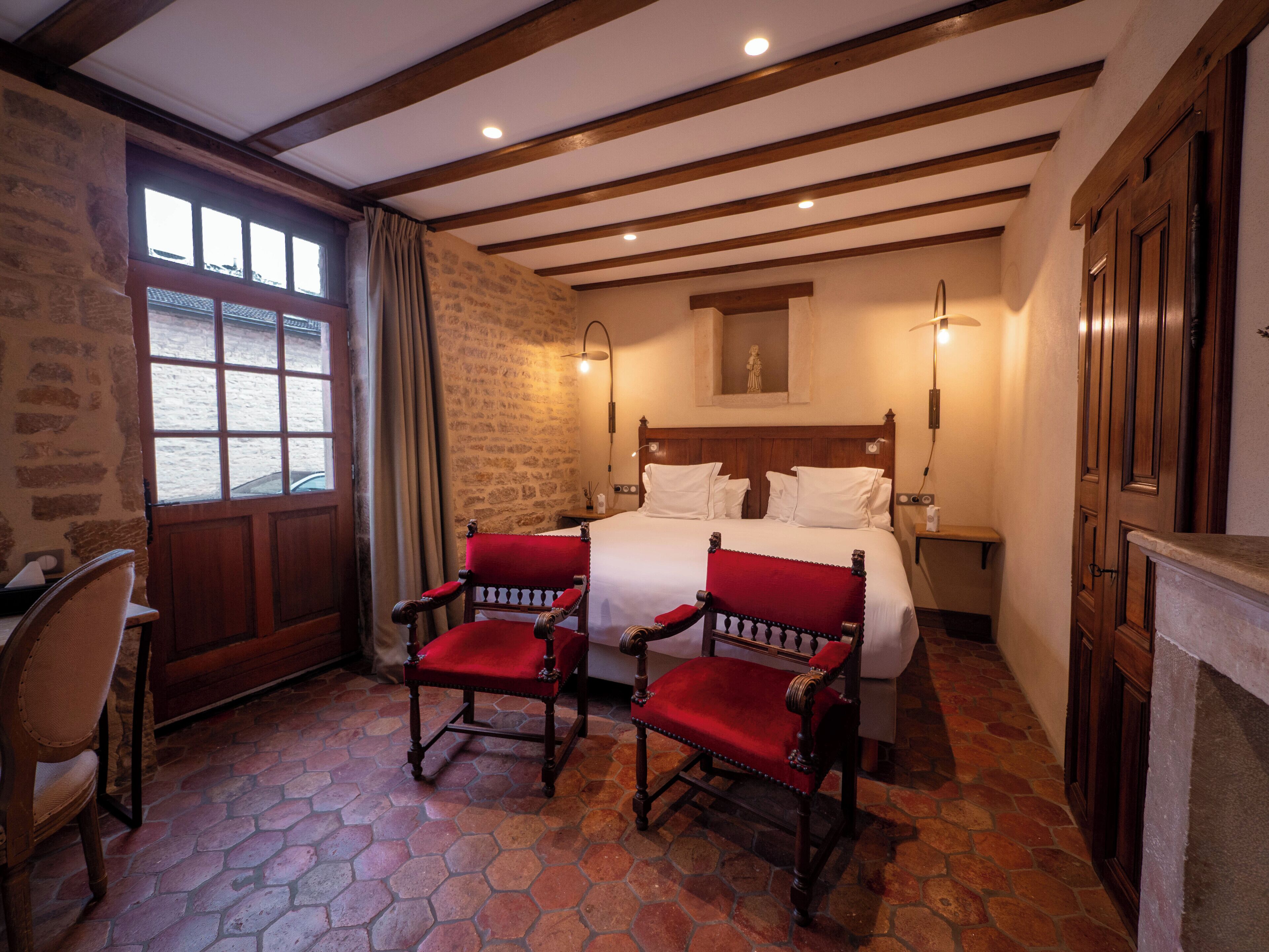 A bedroom with a stone wall, wooden beams, a large bed, two red chairs, and a window with curtains.