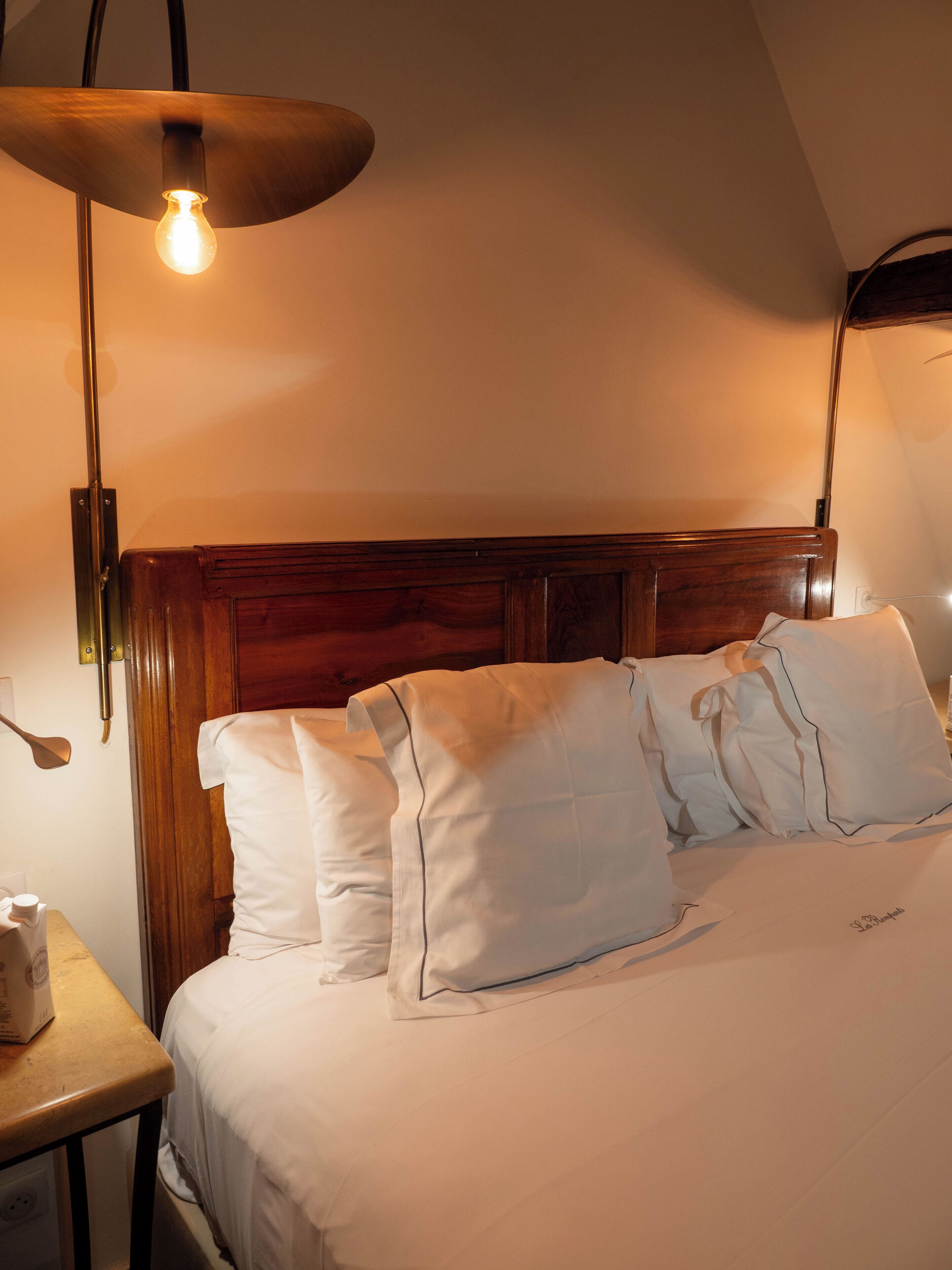 A neatly made bed with white linens and pillows, a wooden headboard, and a single bedside lamp.