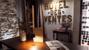 Un hall d'accueil d'hôtel avec une enseigne fixée au mur indiquant « HÔTEL DES VENTES », un comptoir d'accueil en bois, une petite table avec un vase et une bougie, et un mur agrémenté d'une étagère à vin.