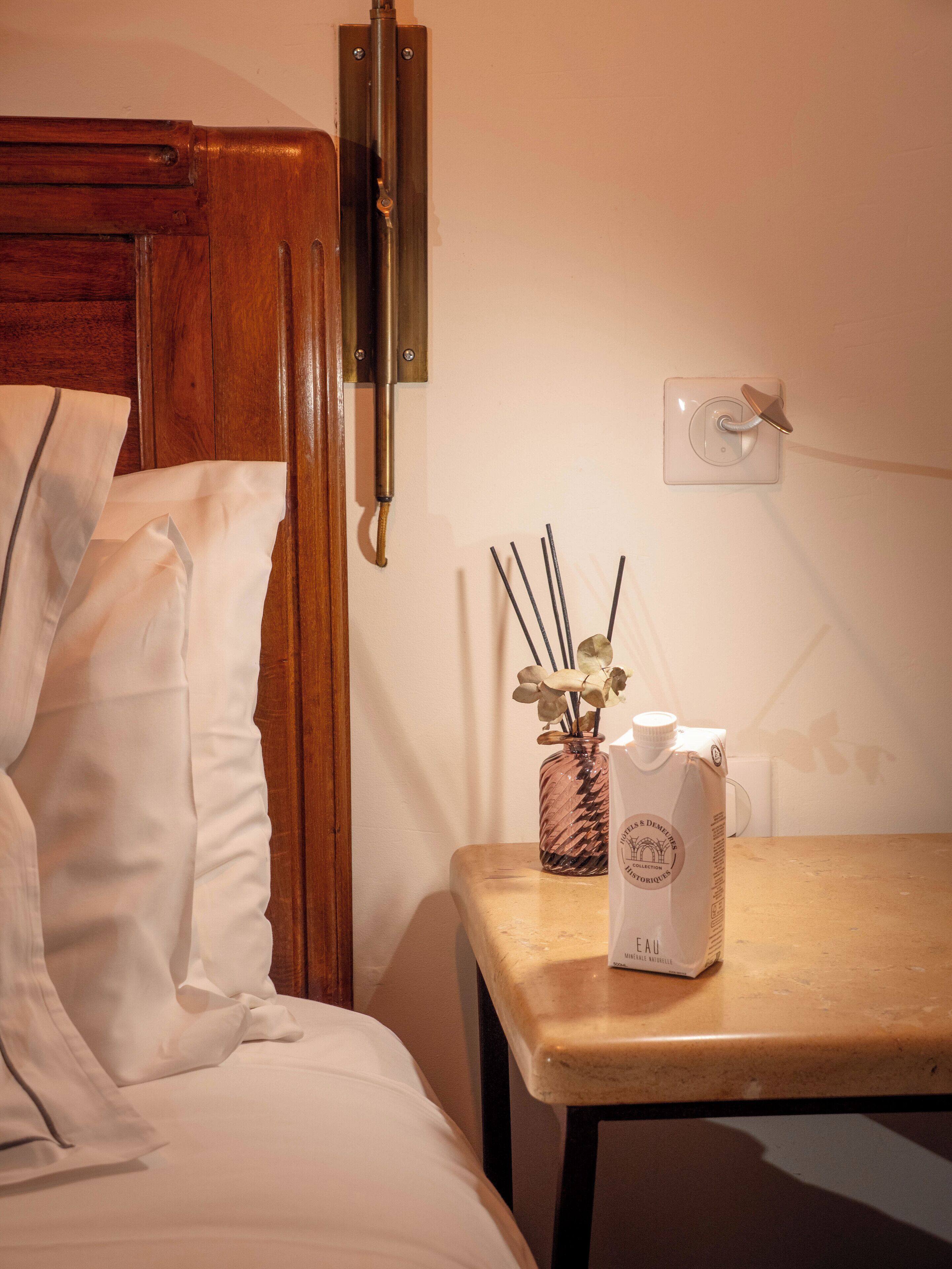 Uma cama bem arrumada com lençóis brancos, uma cabeceira de madeira, uma mesa de cabeceira com um vaso de vidro contendo flores secas e uma garrafa branca com etiqueta.