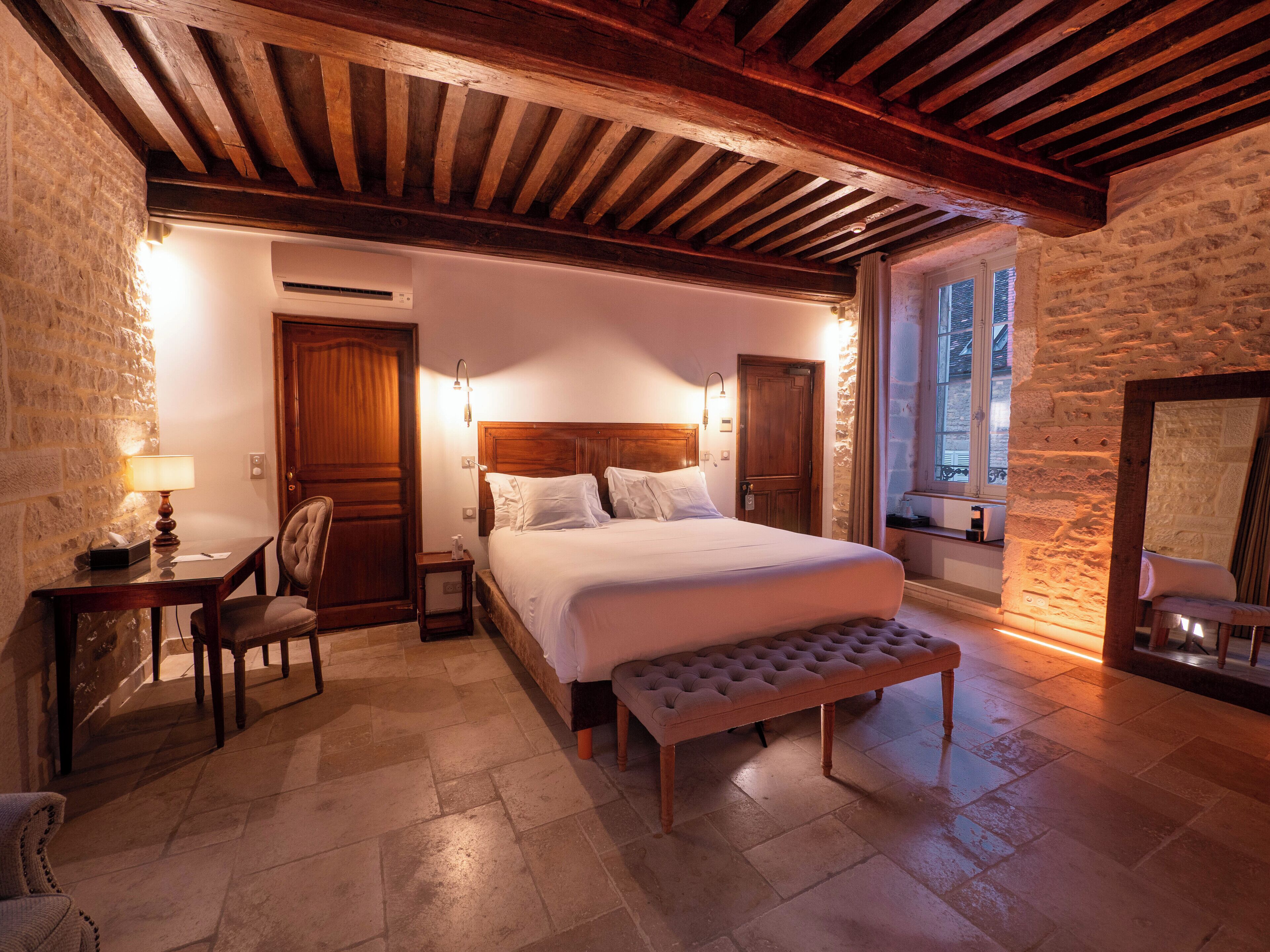 A bedroom with a large bed, a wooden bench, a desk with a lamp, and a stone wall.