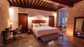 A bedroom with a large bed, a wooden bench, a desk with a lamp, and a stone wall.