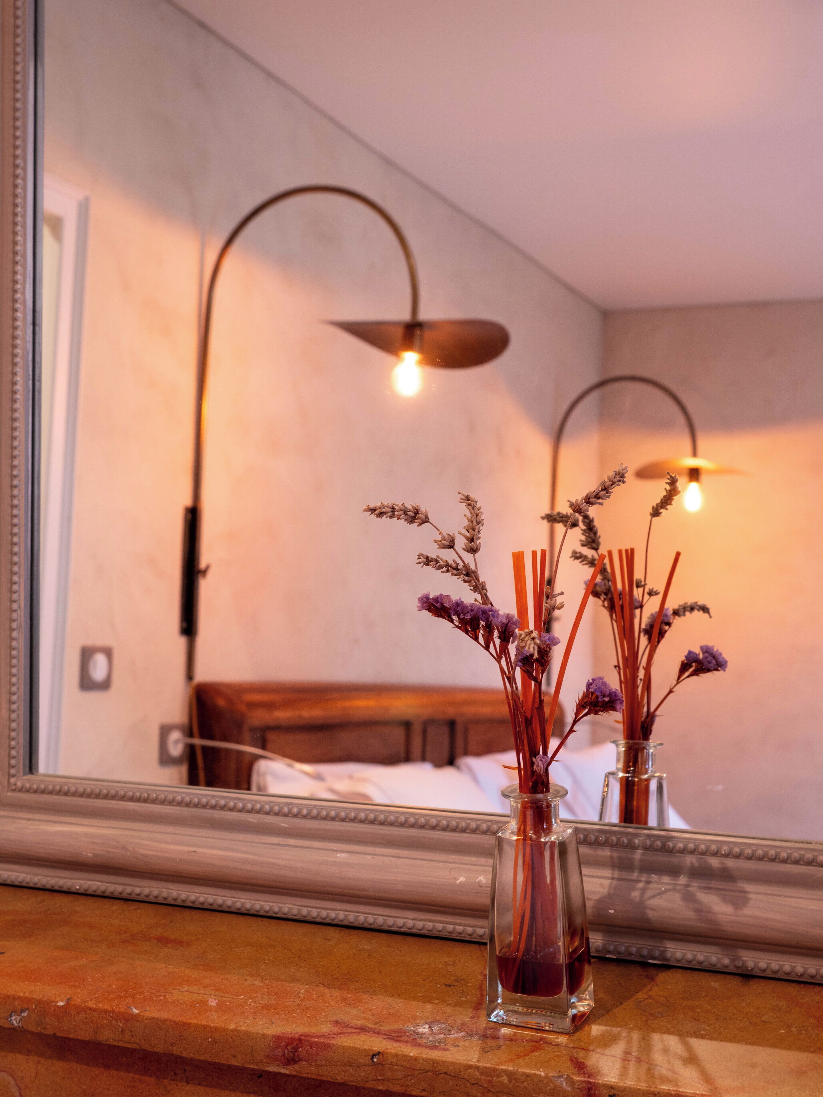 A mirror reflecting a room with a bed, a vase with dried flowers, and a wall-mounted lamp.