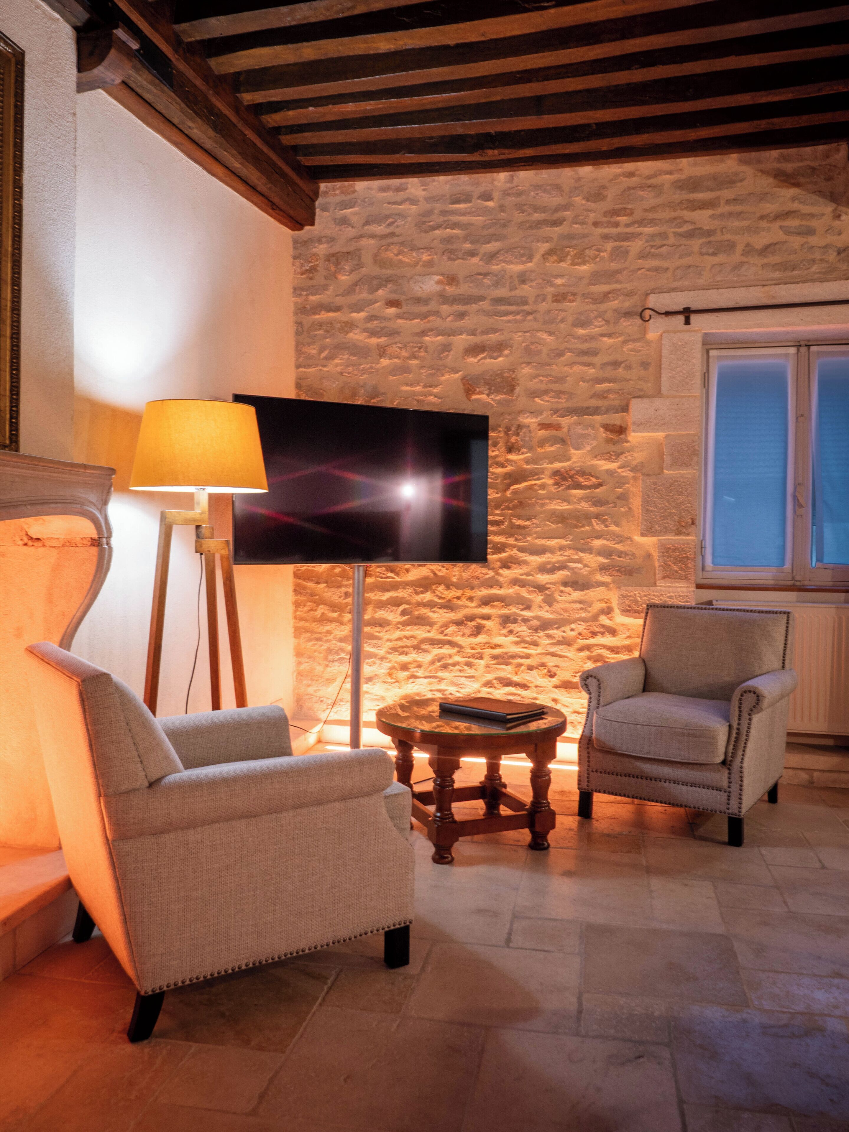 Une pièce chaleureuse avec un mur en pierre, un téléviseur à écran plat, deux fauteuils, une petite table ronde et un lampadaire.