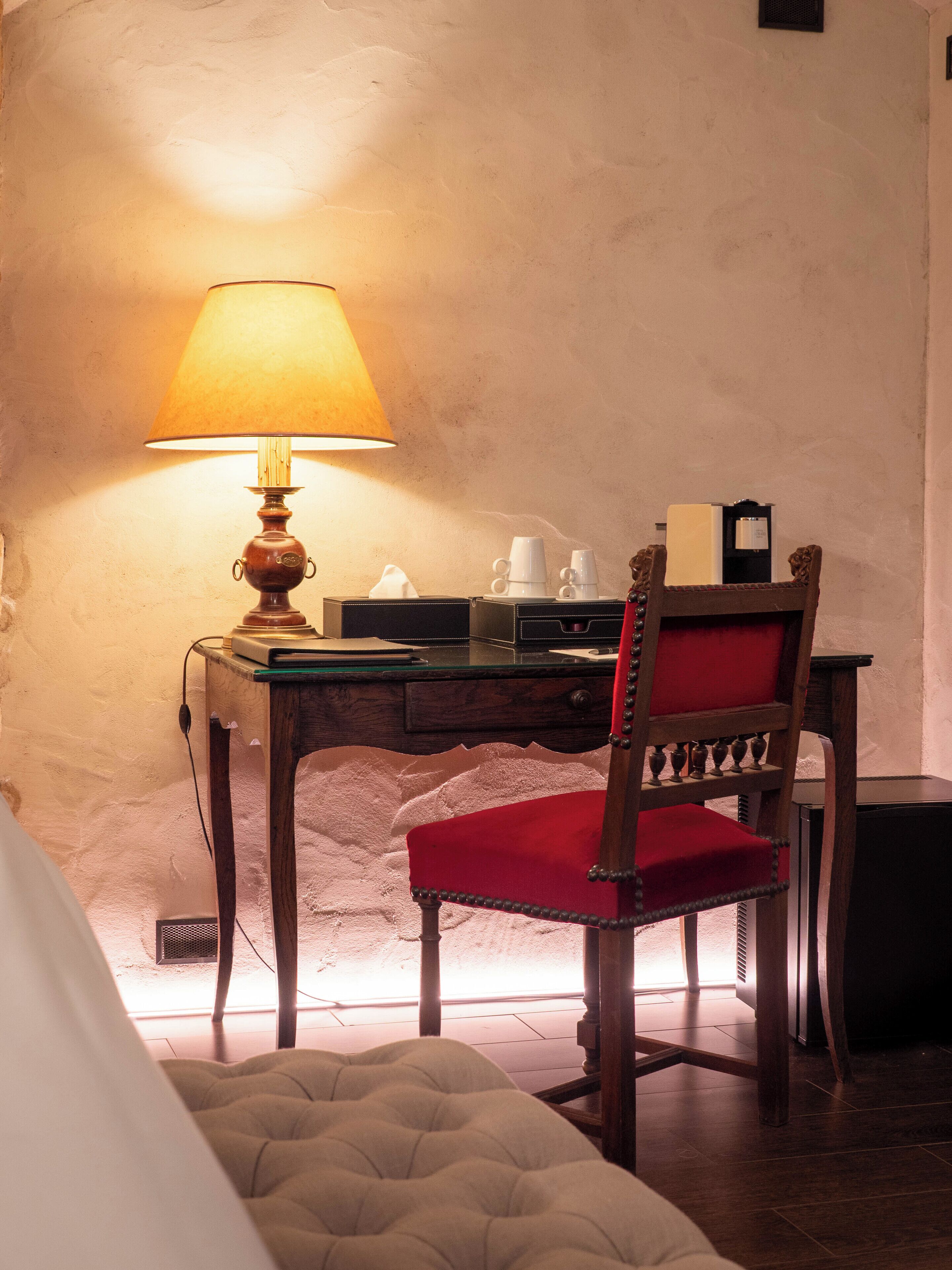 Une pièce avec un bureau en bois, une chaise rembourrée de tissu rouge, une lampe et un lit recouvert d’une couette blanche.