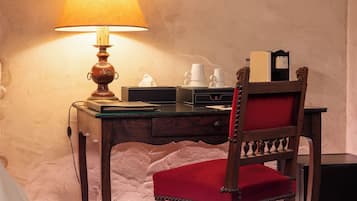 Une pièce avec un bureau en bois, une chaise rembourrée de tissu rouge, une lampe et un lit recouvert d’une couette blanche.