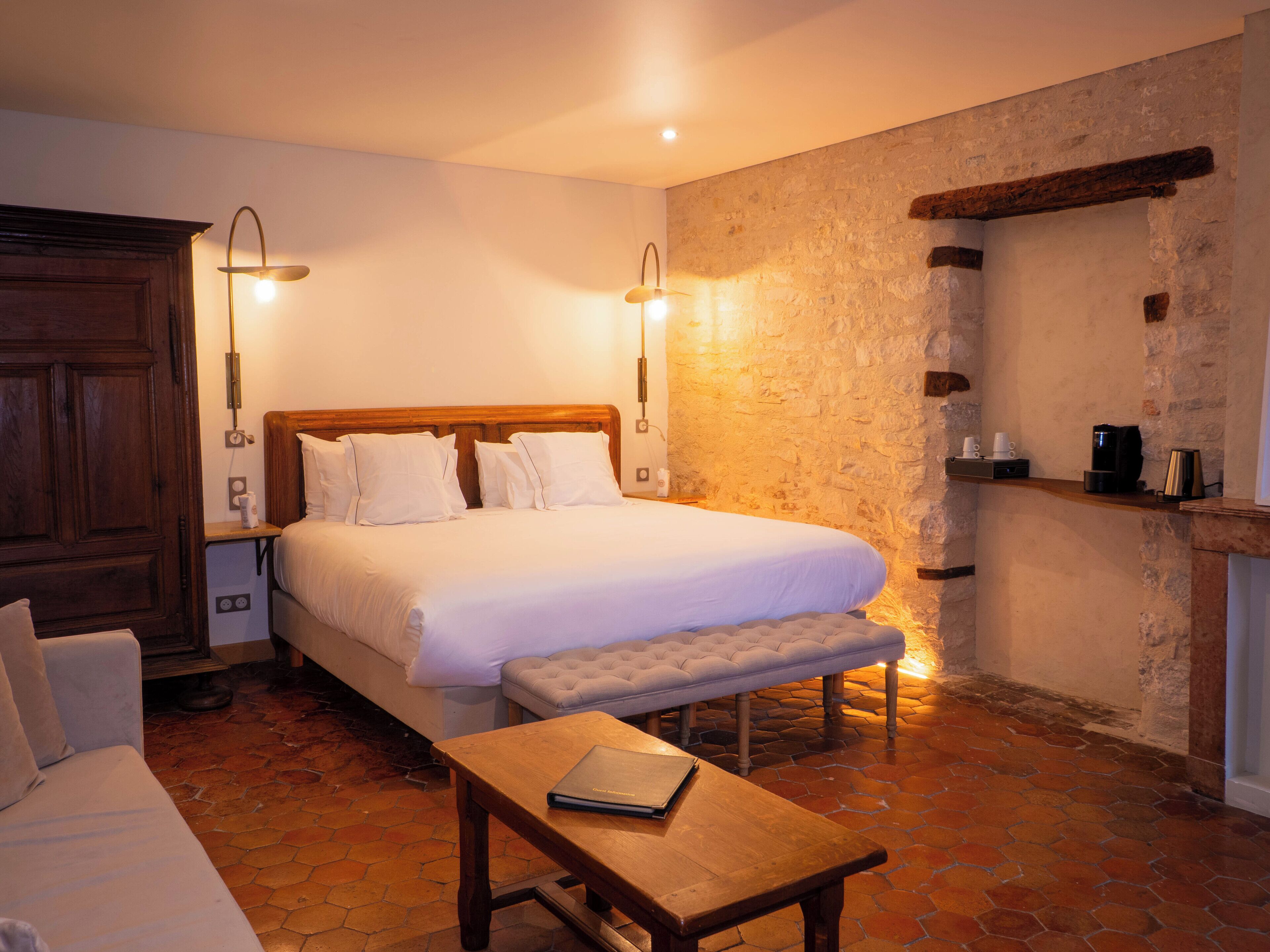 A bedroom with a wooden bed, bedside lamps, a bench, a small table, and a stone wall.