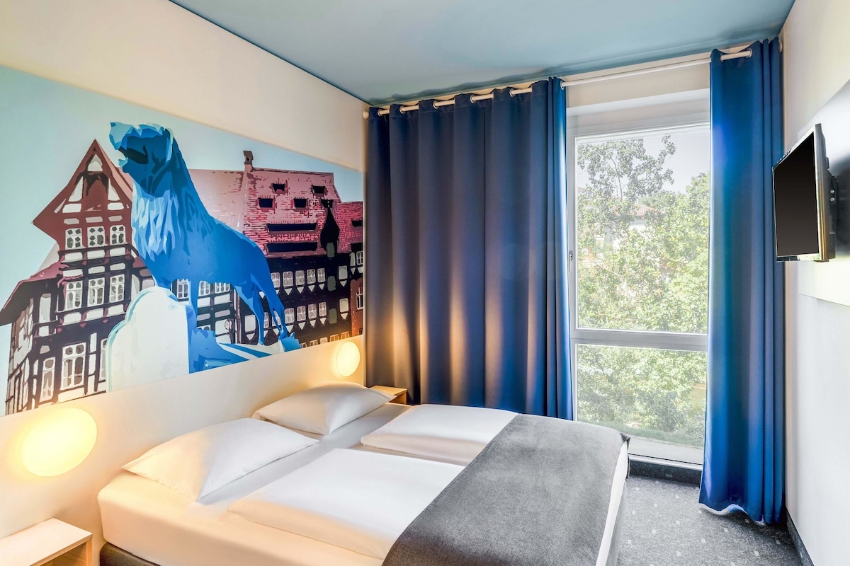 A hotel room with a bed, a wall mural, a window with curtains, and a flat-screen TV.