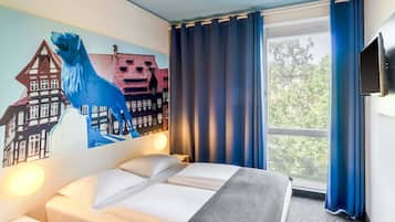 A hotel room with a bed, a wall mural, a window with curtains, and a flat-screen TV.