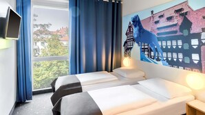 Habitación de hotel con dos camas, un ventanal con cortinas, un televisor montado y un mural con un caballo y edificios en la pared.