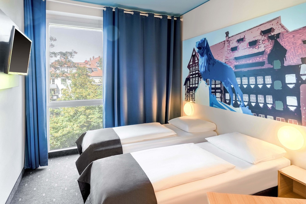 A hotel room with two beds, a large window with curtains, a mounted TV, and a mural of a horse and buildings on the wall.