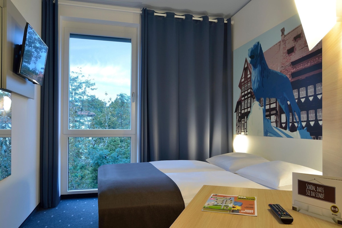 A hotel room with a bed, a desk with a remote control, a magazine, and a sign in German.