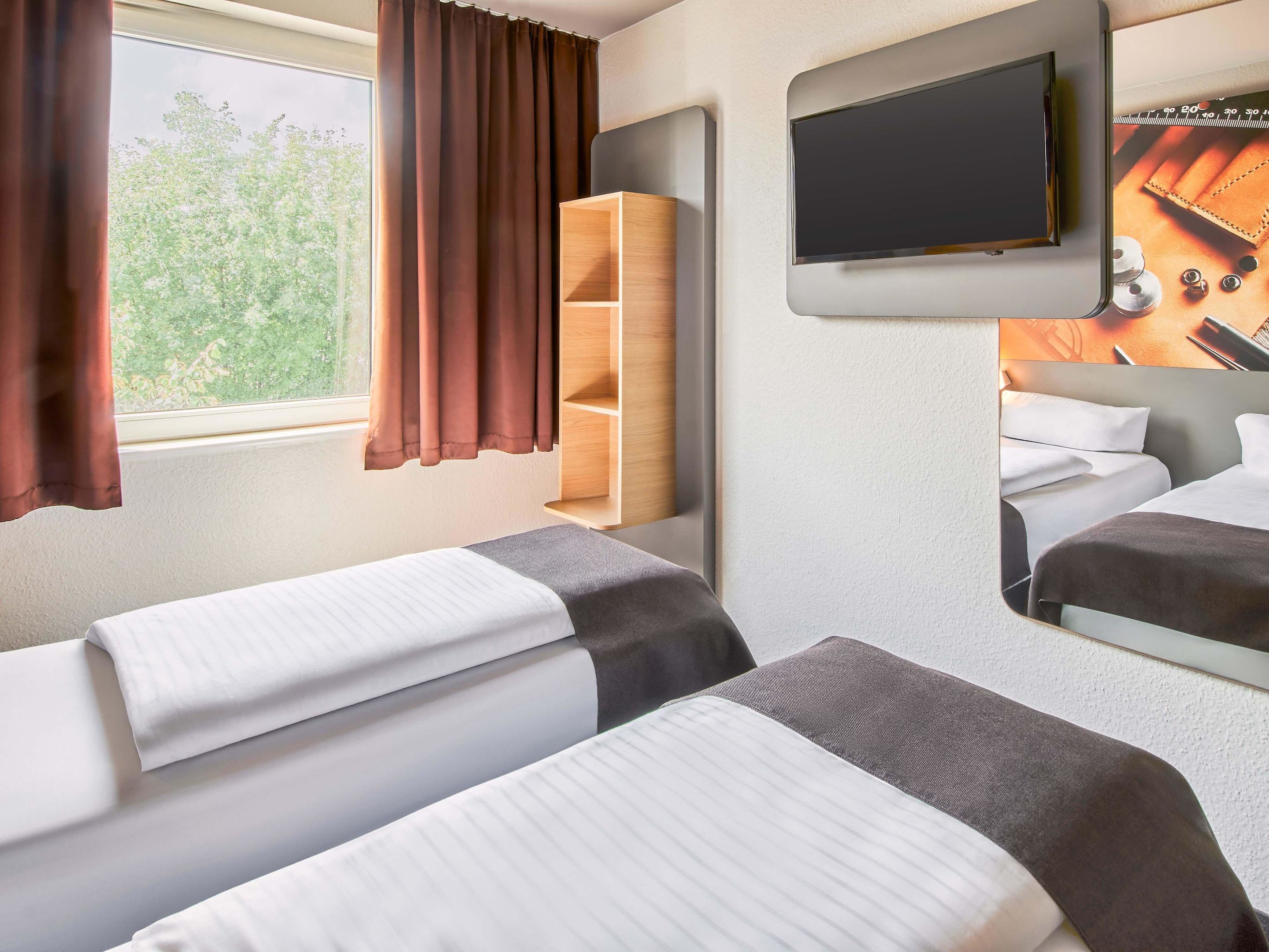 A hotel room with two beds, a TV mounted on the wall, and a window with curtains.