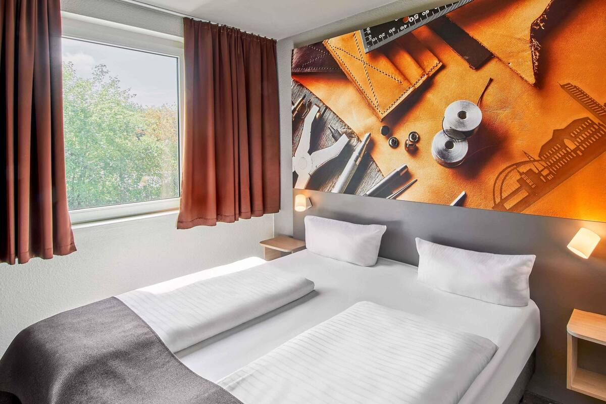 Ein Hotelzimmer mit einem großen Bett, einem Fenster mit Vorhängen, einem Kopfteil mit Stadtansicht-Wandgemälde und einem Nachttisch mit Lampe.