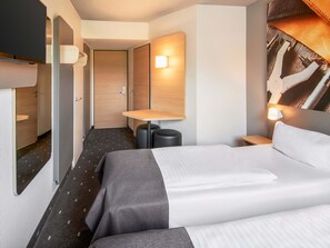 Une chambre d’hôtel comprenant un lit, des tables de chevet, un bureau, une chaise, une lampe et un téléviseur fixé au mur.