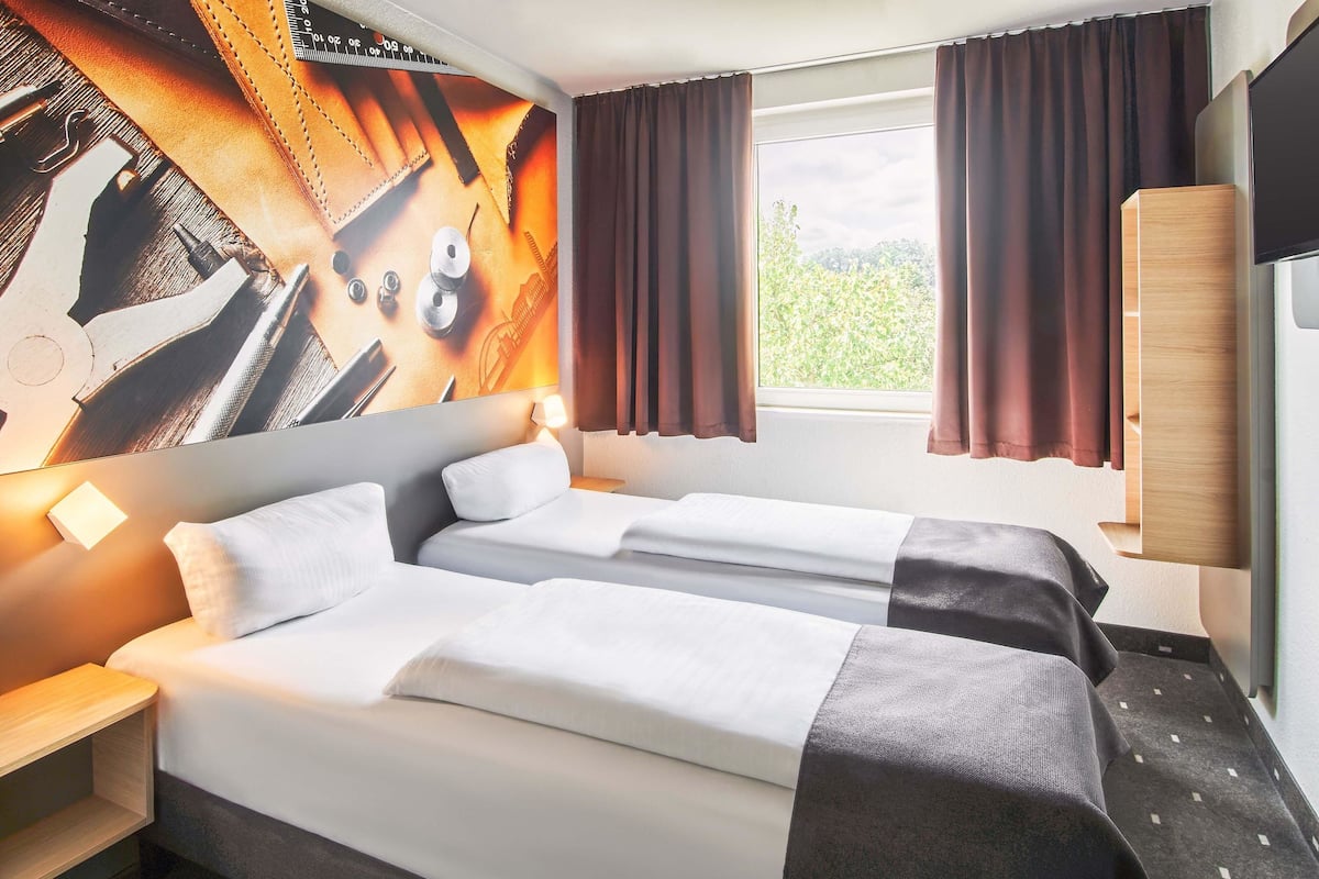 Ein Hotelzimmer mit zwei Betten, einem großen Fenster mit Vorhängen, einem an der Wand montierten Fernseher und einem Holzschrank.