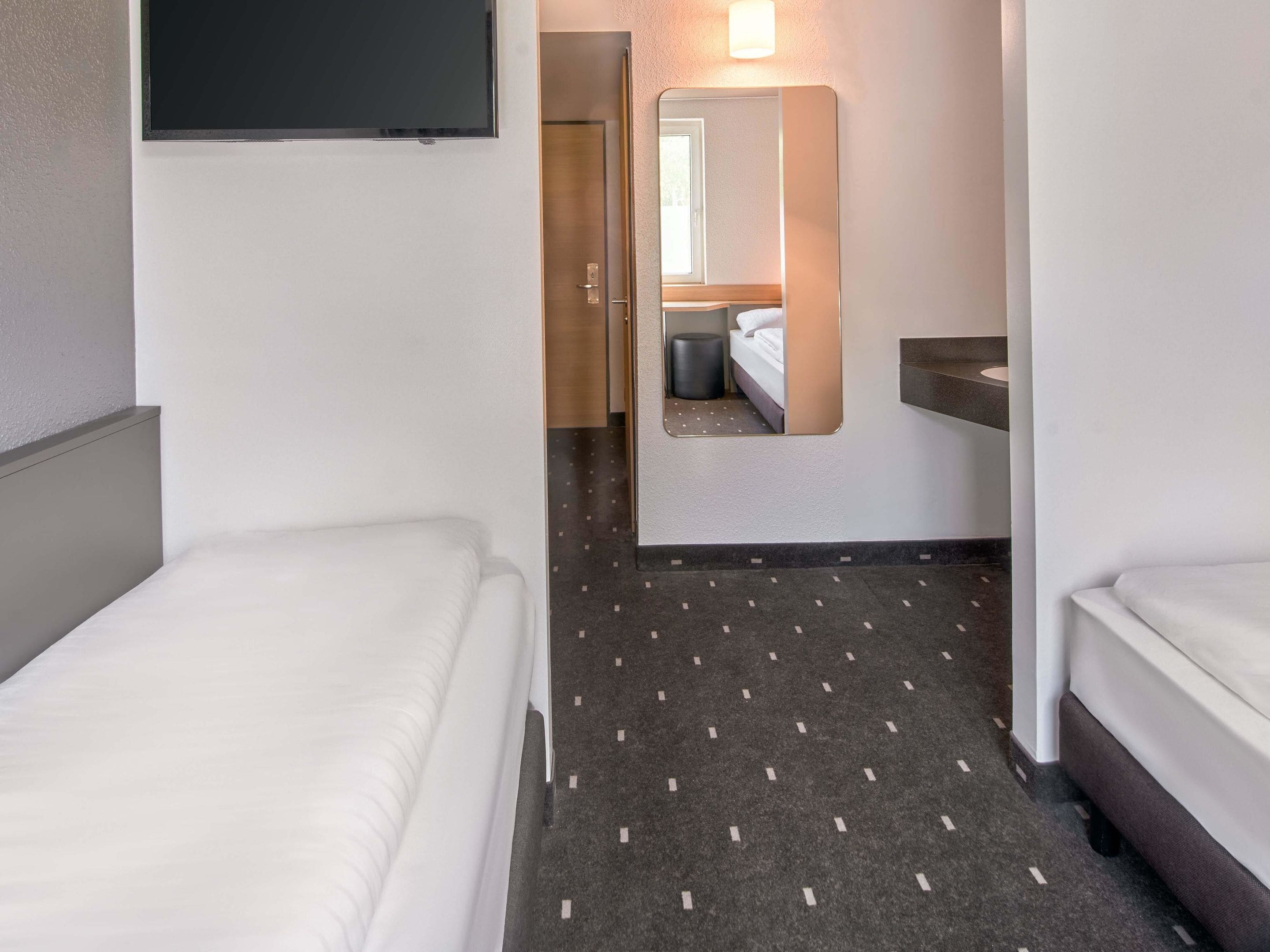 Habitación de hotel con dos camas, televisión, espejo y baño visible a través de una puerta abierta.