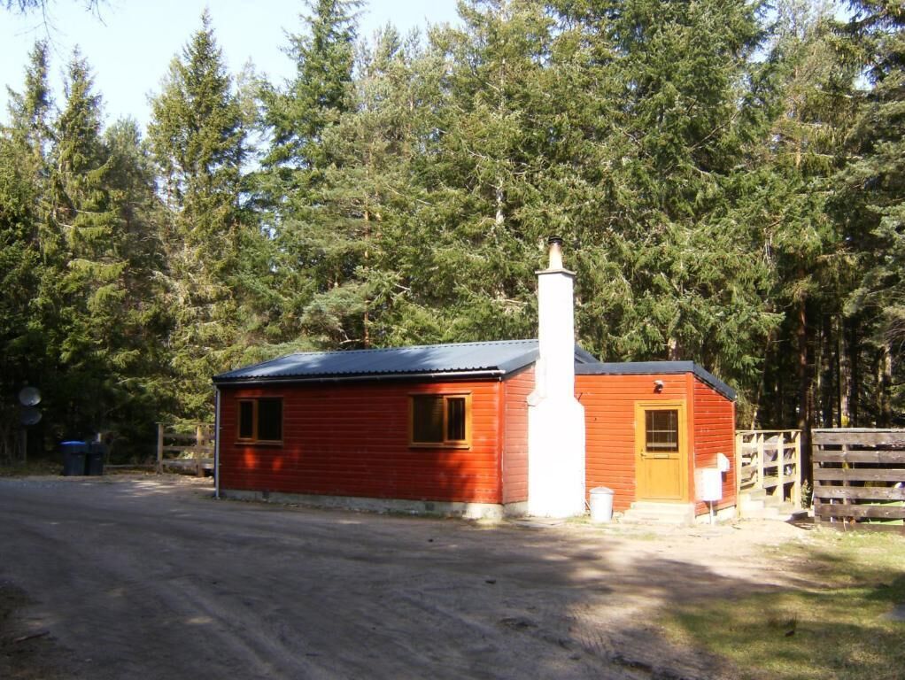 A red wooden cabin with a chimney, a fenced yard, and a forested backdrop.