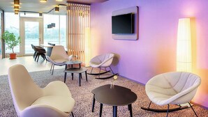 A modern lounge area with contemporary furniture, a flat-screen TV, and a view through large windows. - B&B Hotel Bamberg (Bamberg)