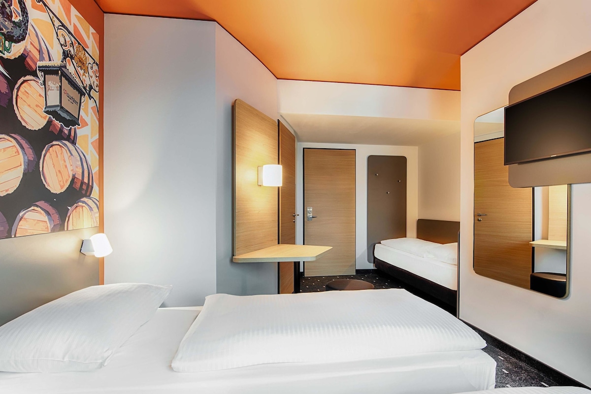 Una habitación de hotel moderna con cama, una mesita pequeña, televisor de pared y una puerta que da a otra habitación.