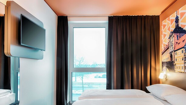 Habitación de hotel con cama, televisor en la pared, ventana con cortinas y vista a un edificio.