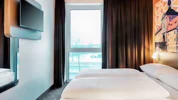 Ein Hotelzimmer mit Bett, Wandfernseher, Fenster mit Vorhängen und Blick auf ein Gebäude.