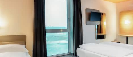A hotel room with two beds, a large window with curtains, a flat-screen TV, and a small table.