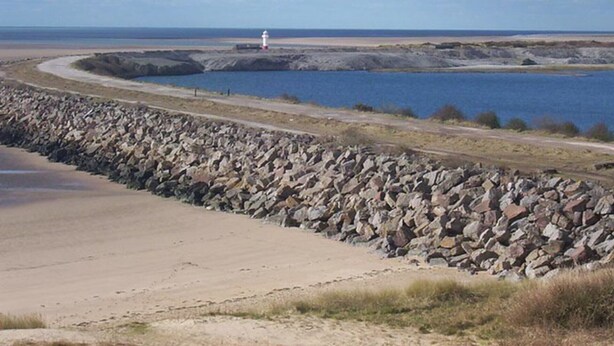 Een kustlandschap met een rotsachtige zeedijk, een zandstrand en een vuurtoren in de verte.
