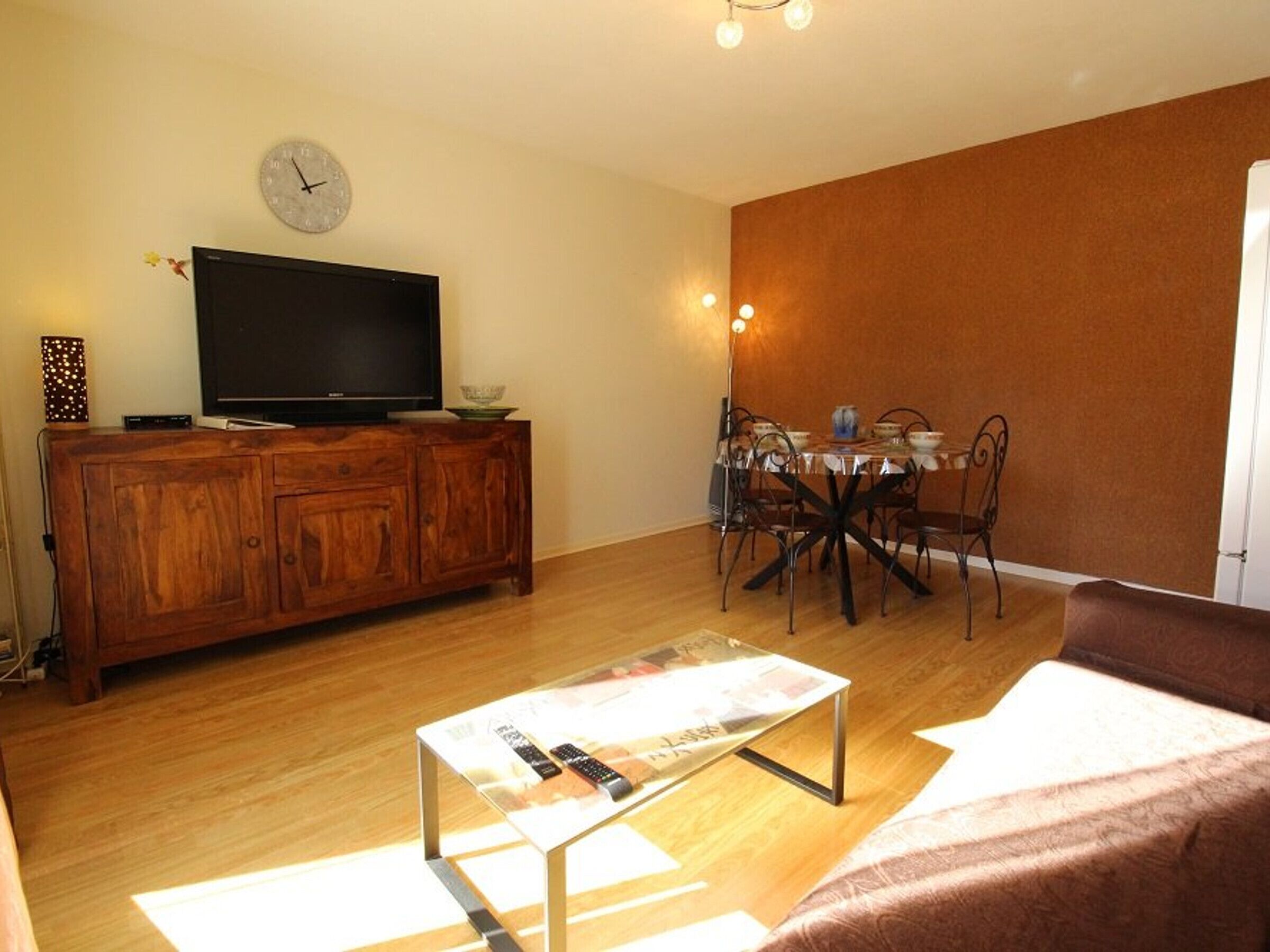 A living room with a wooden cabinet, a flat-screen TV, a dining table with chairs, and a magazine rack.