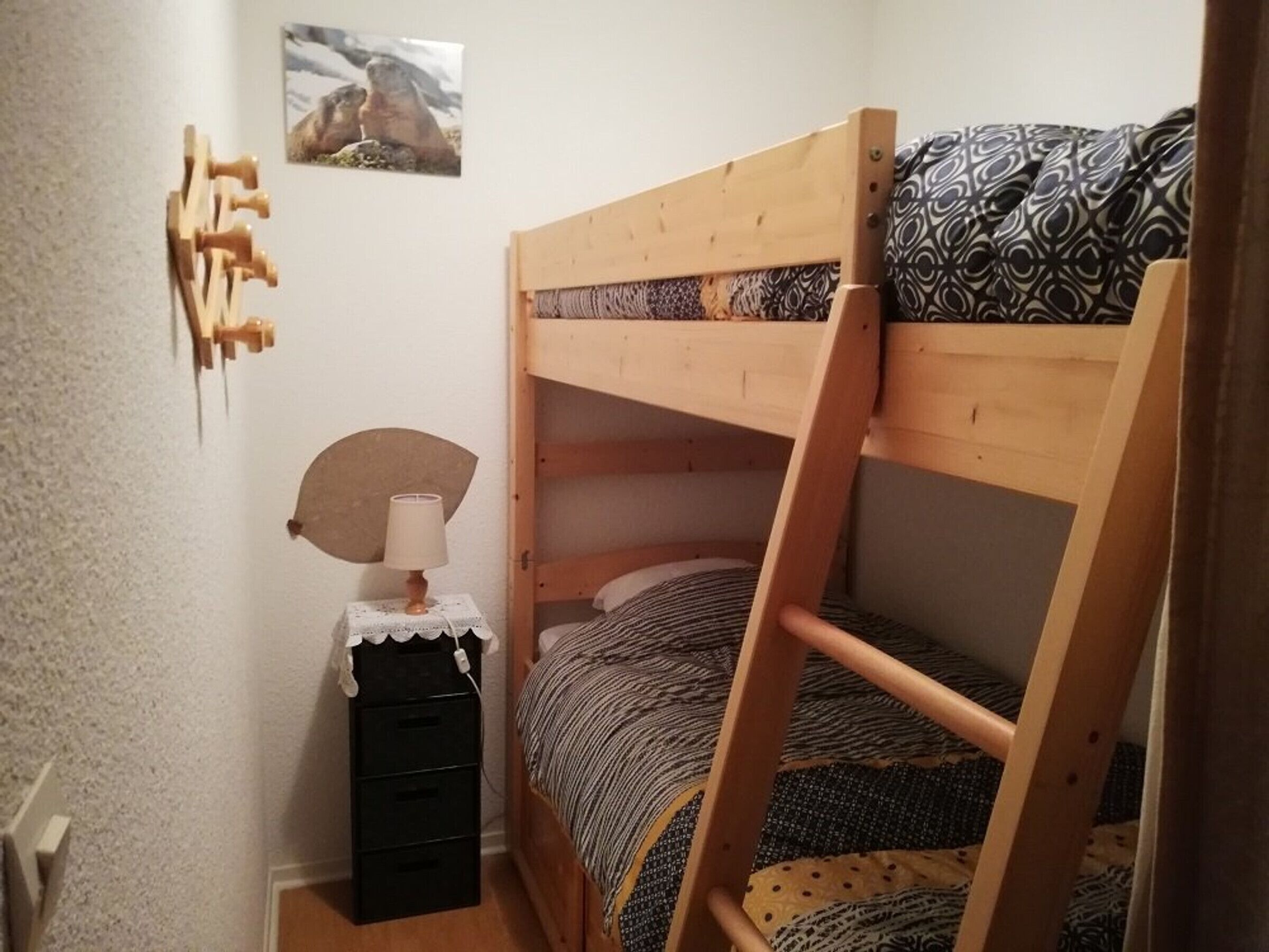 Een stapelbed met houten frame, een nachtkastje met lamp en een klein opbergmeubel.