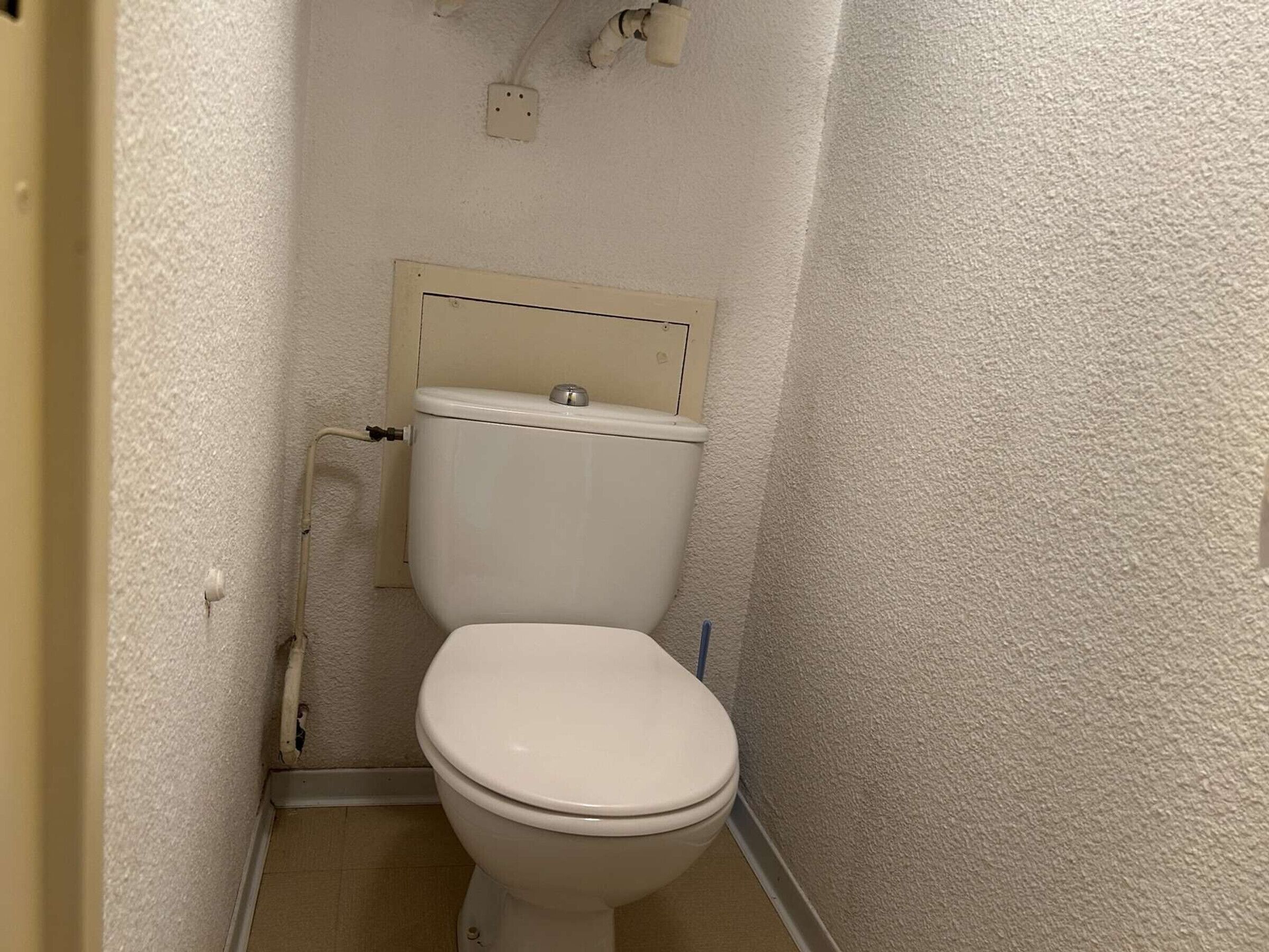 Une petite salle de bain étroite, avec des toilettes blanches, une tuyauterie apparente et une prise électrique fixée au mur.