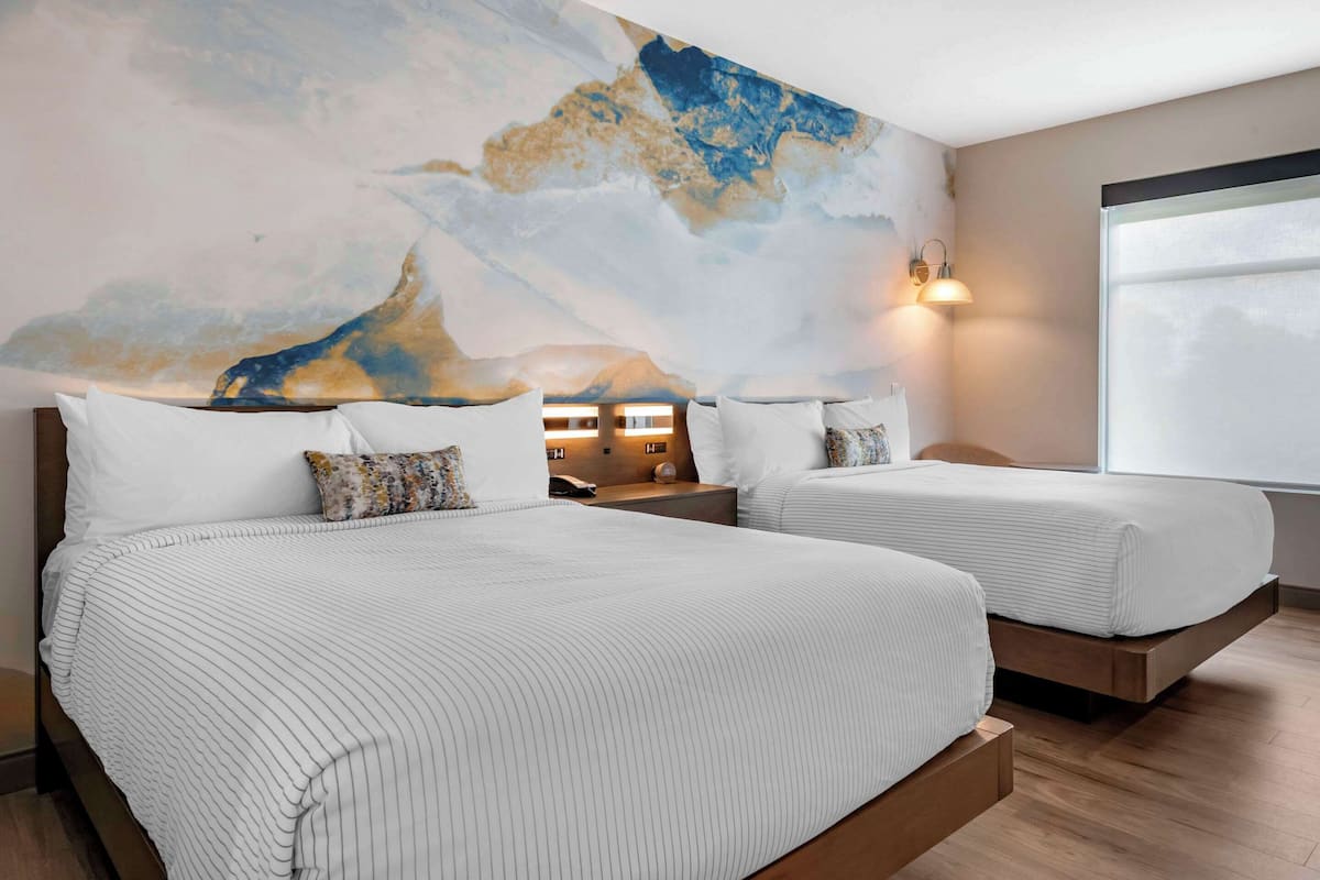 A hotel room with two beds, a large abstract mural on the wall, a bedside table with a lamp, and a window with a view.